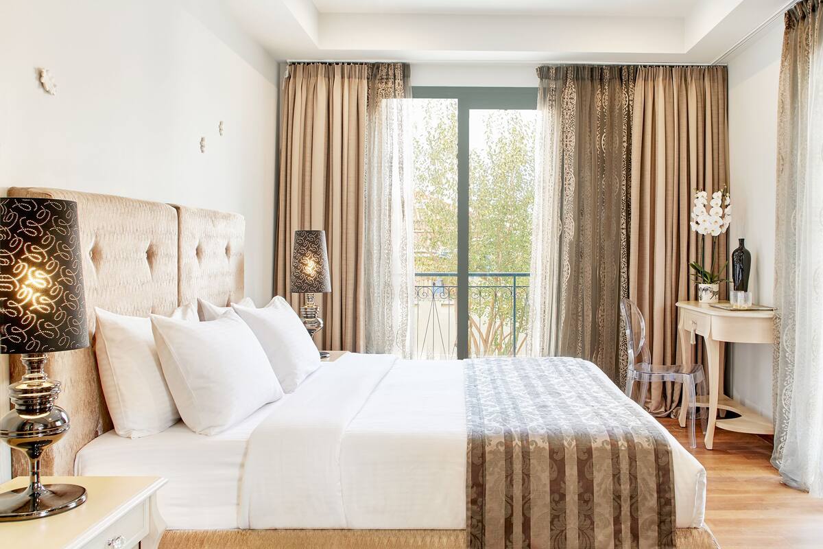 A bedroom with a large bed, a bedside table with a lamp, a desk with a chair, and a view of the outdoors through curtains.