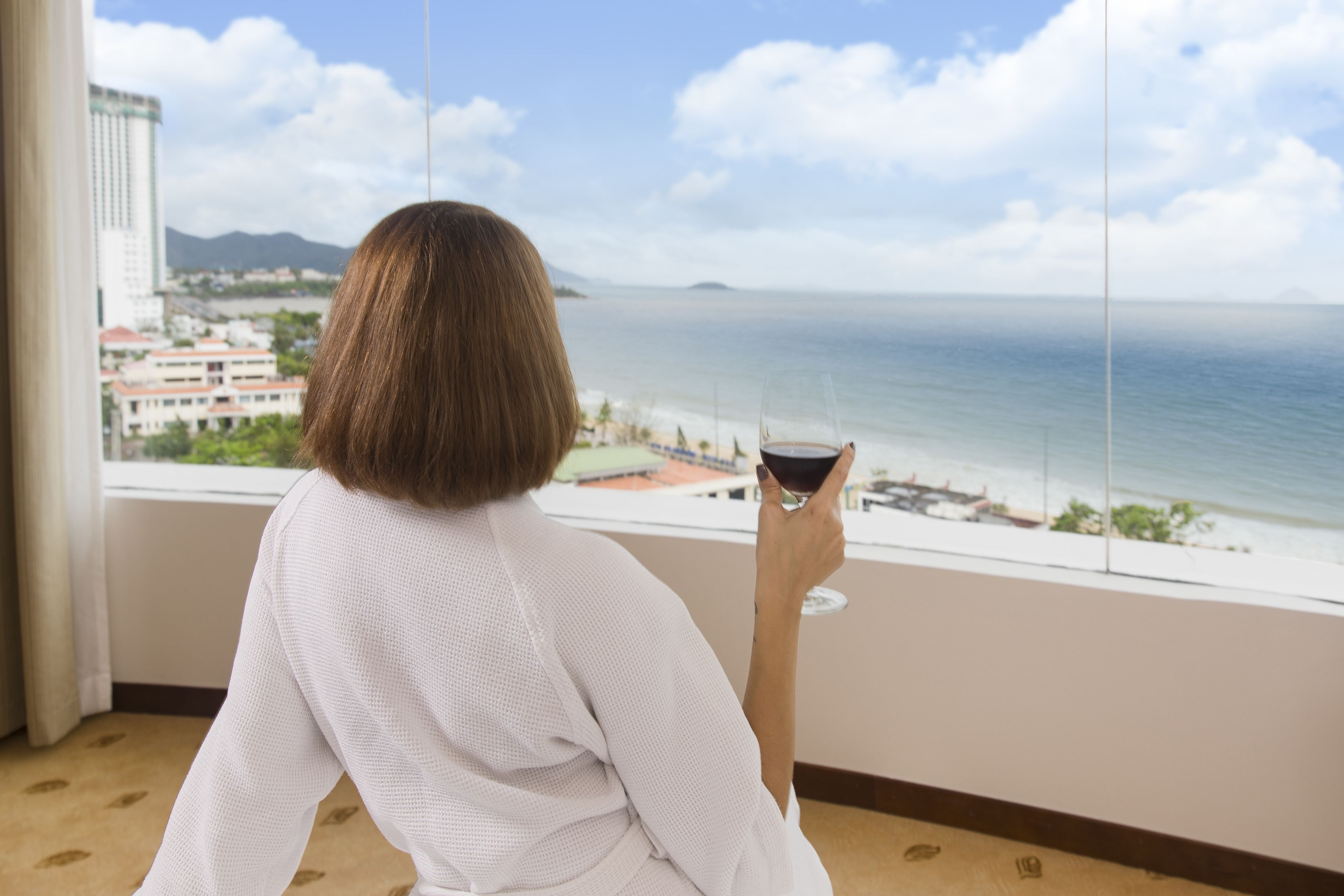 Une personne assise près d’une fenêtre, avec vue sur la mer et les bâtiments.