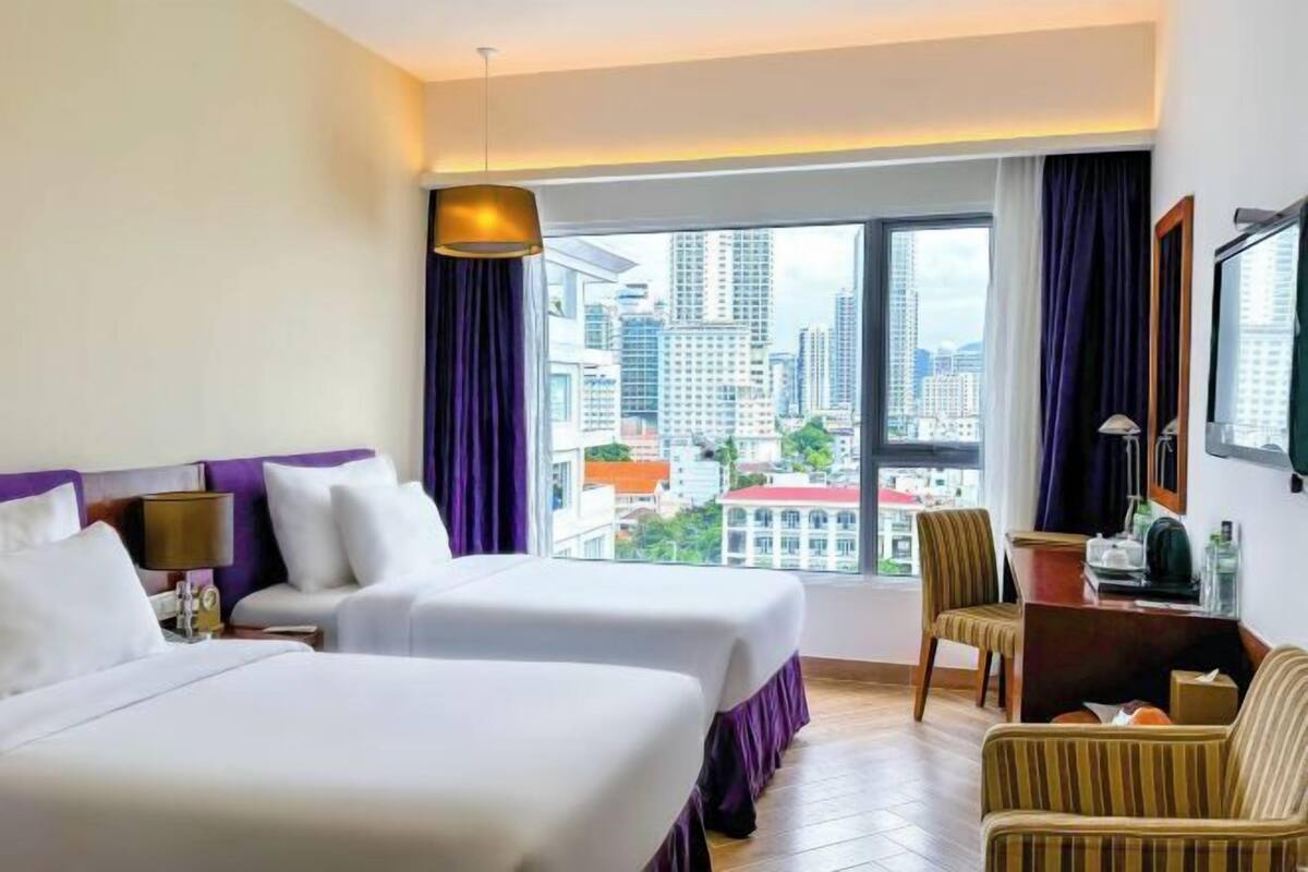 Habitación de hotel con dos camas, un escritorio, una silla y un amplio ventanal con vistas a la ciudad.