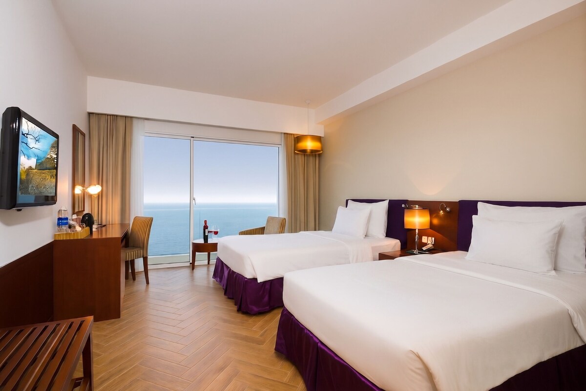 A hotel room with two beds, a desk, a chair, a TV, and a view of the sea.