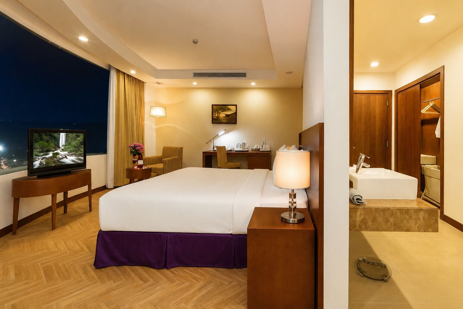 A hotel room with a bed, a TV, a desk, a chair, a lamp, a painting, a bathroom with a sink and mirror.