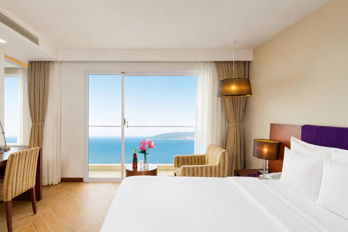 A hotel room with a bed, a desk, a chair, a lamp, and a view of the sea.