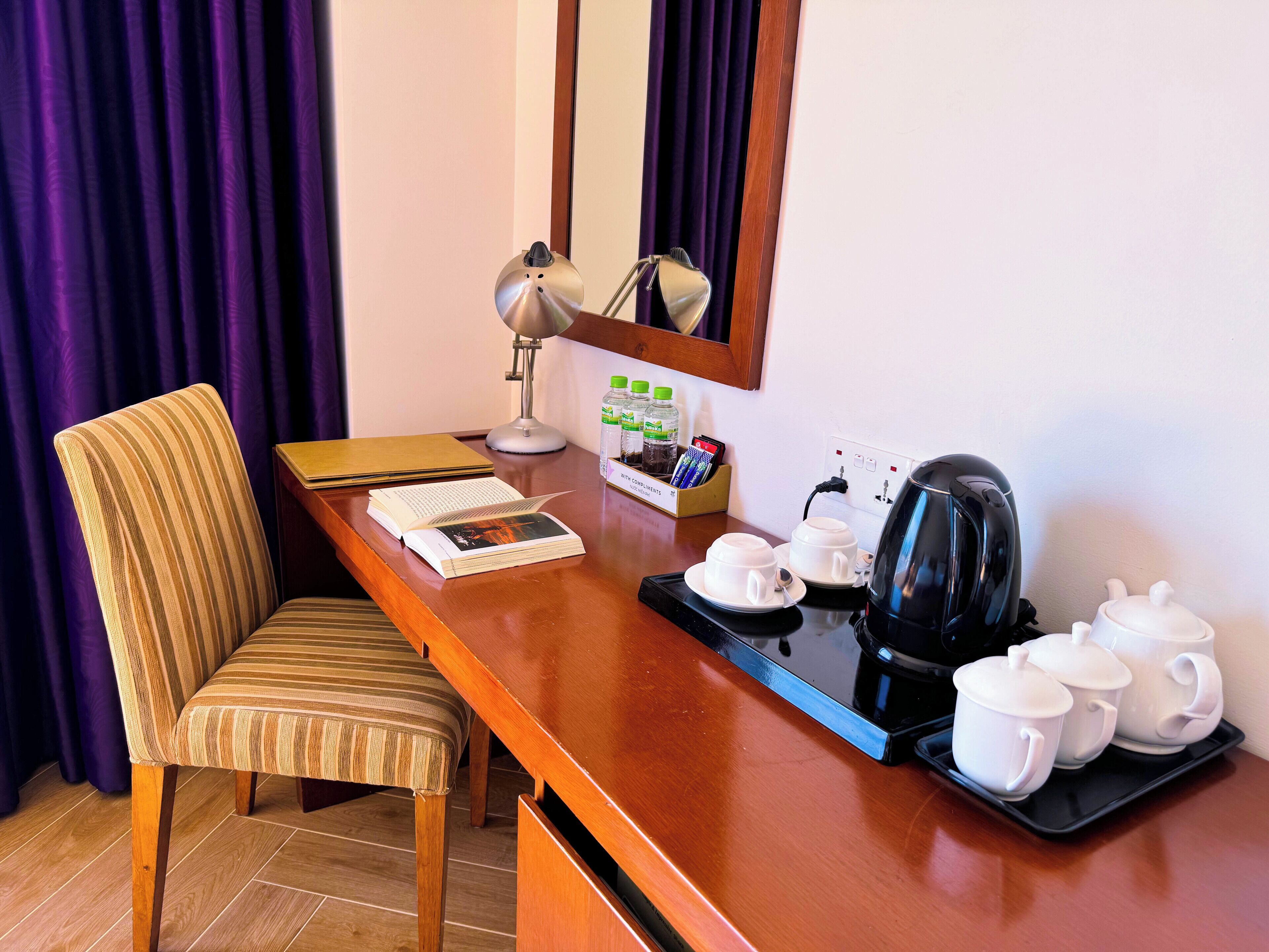 A hotel room with a wooden desk, a striped chair, a lamp, a coffee maker, and a tray with cups and a teapot.