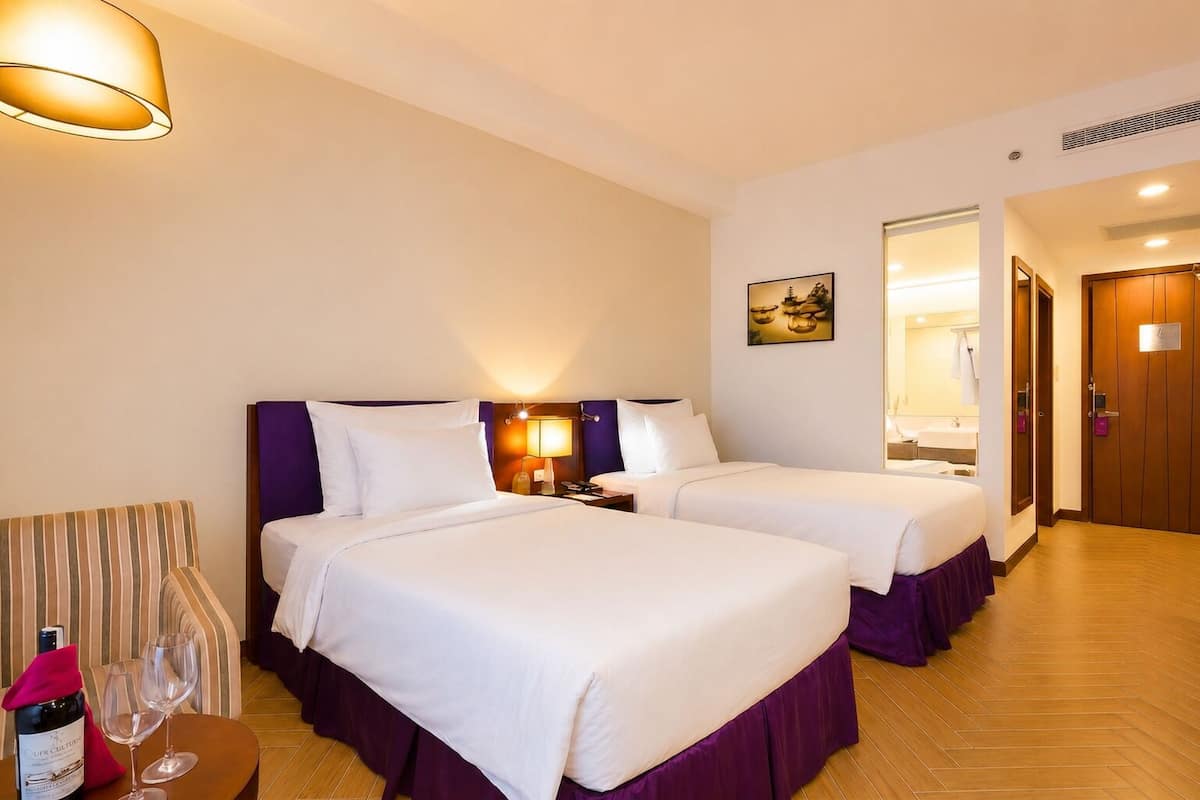 Habitación de hotel con dos camas, una mesita de noche, una lámpara, una botella de vino y copas sobre una pequeña mesa.