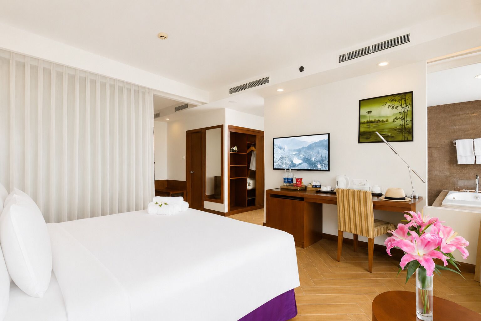 A hotel room with a bed, a desk, a chair, a TV, a bathroom with a bathtub, and a vase with flowers.
