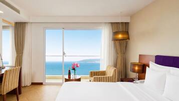 A hotel room with a bed, a desk, a chair, a lamp, and a view of the sea.