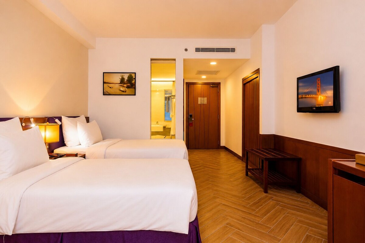 A hotel room with two beds, a television, a wooden bench, and a hallway leading to a bathroom.