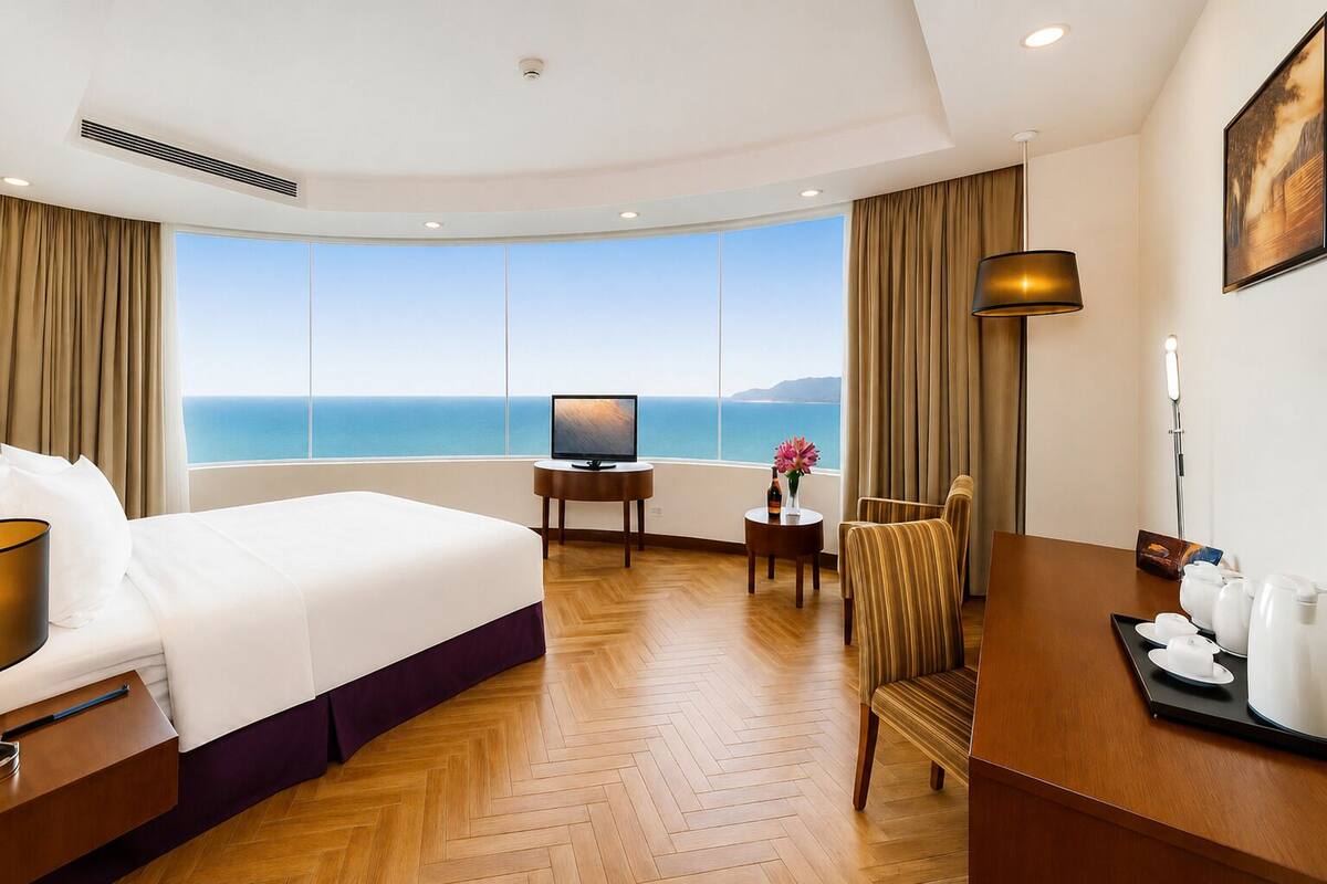 Habitación de hotel con una cama grande, televisión, una mesita con una botella y flores, una silla y vistas al mar.