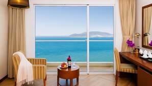 Una camera d'hotel con un'ampia porta finestra vista mare, una scrivania in legno con un vaso di fiori, una sedia e un tavolino con una bottiglia e un bicchiere.