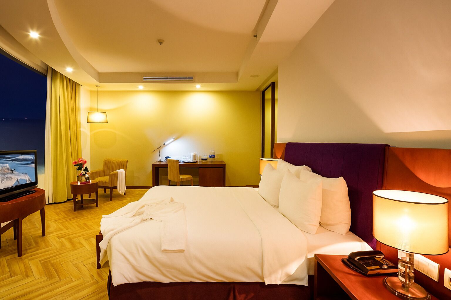 Quarto de hotel com cama, mesa de cabeceira, luminária, televisão, escrivaninha, cadeira e vista para o exterior.