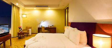 Quarto de hotel com cama, mesa de cabeceira, luminária, televisão, escrivaninha, cadeira e vista para o exterior.