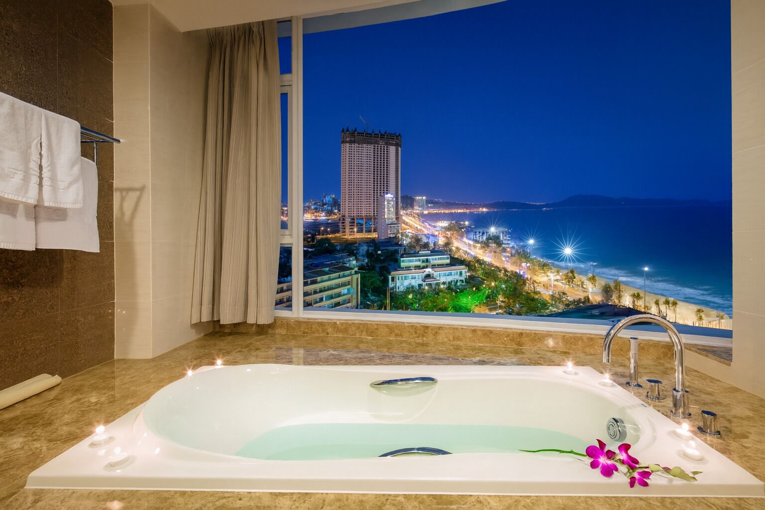 A modern bathroom with a large bathtub, a view of a cityscape and coastline at night.