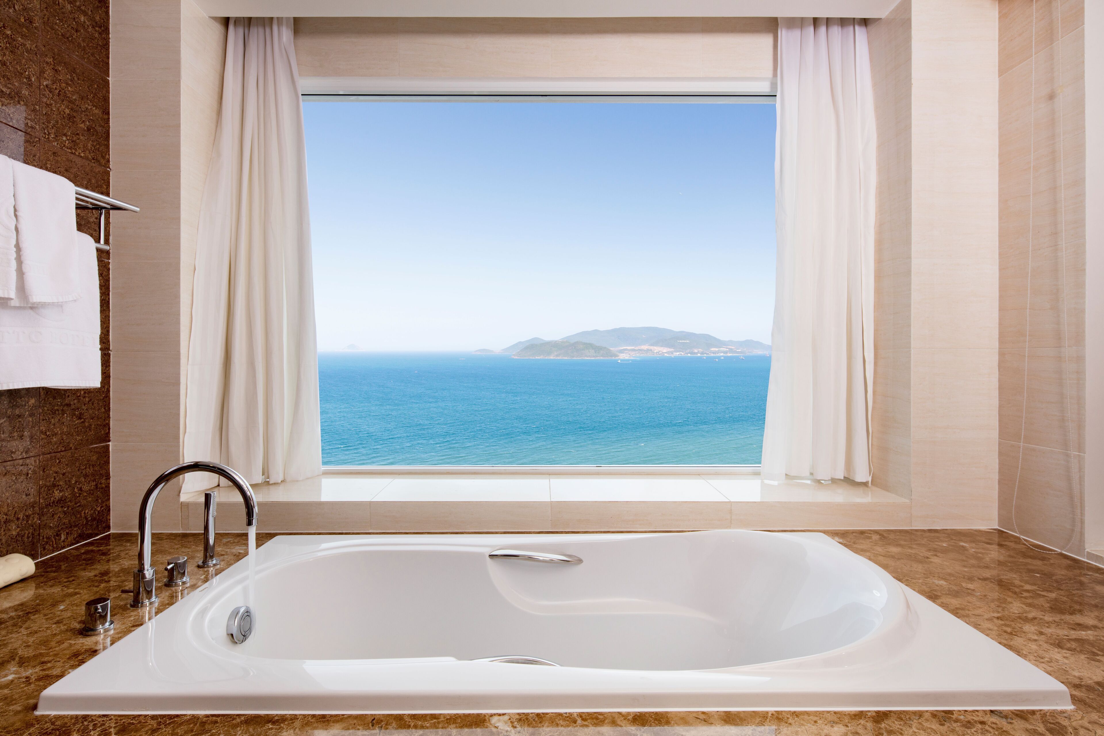 A modern bathroom with a large bathtub, a window with a view of the sea, and marble flooring.