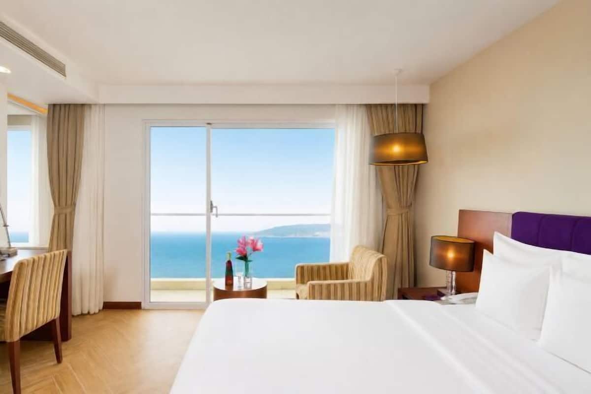 Habitación de hotel con cama, escritorio, silla, mesita de noche, lámpara y vista al mar.