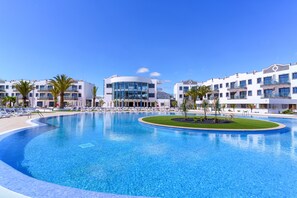 Una amplia piscina rodeada de edificios modernos con balcones, palmeras y tumbonas.
