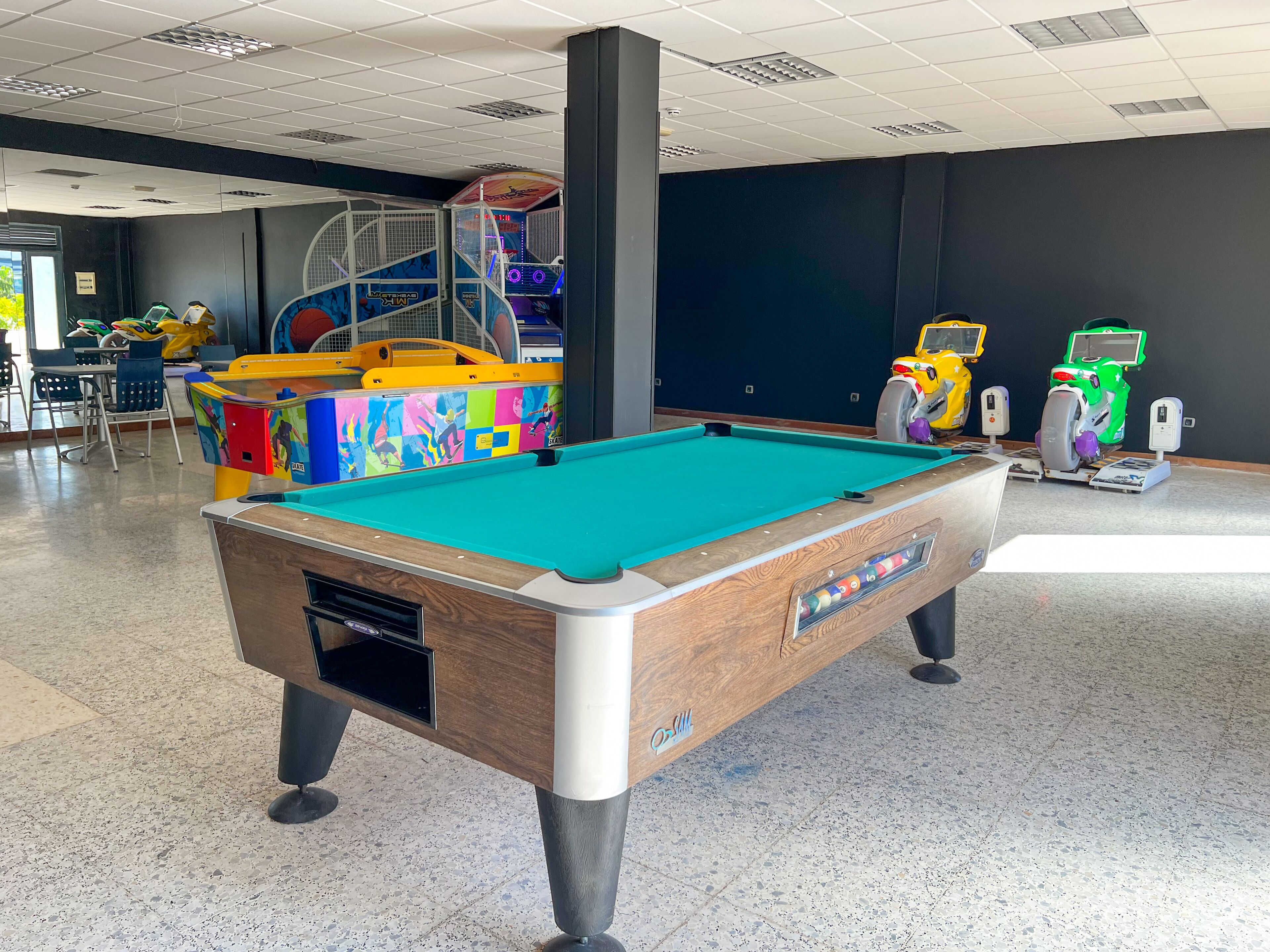 Una mesa de billar en una sala con una estructura en el techo, un área de juegos infantiles y una zona de estar con sillas.
