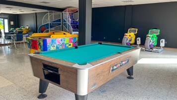 Una mesa de billar en una sala con una estructura en el techo, un área de juegos infantiles y una zona de estar con sillas.