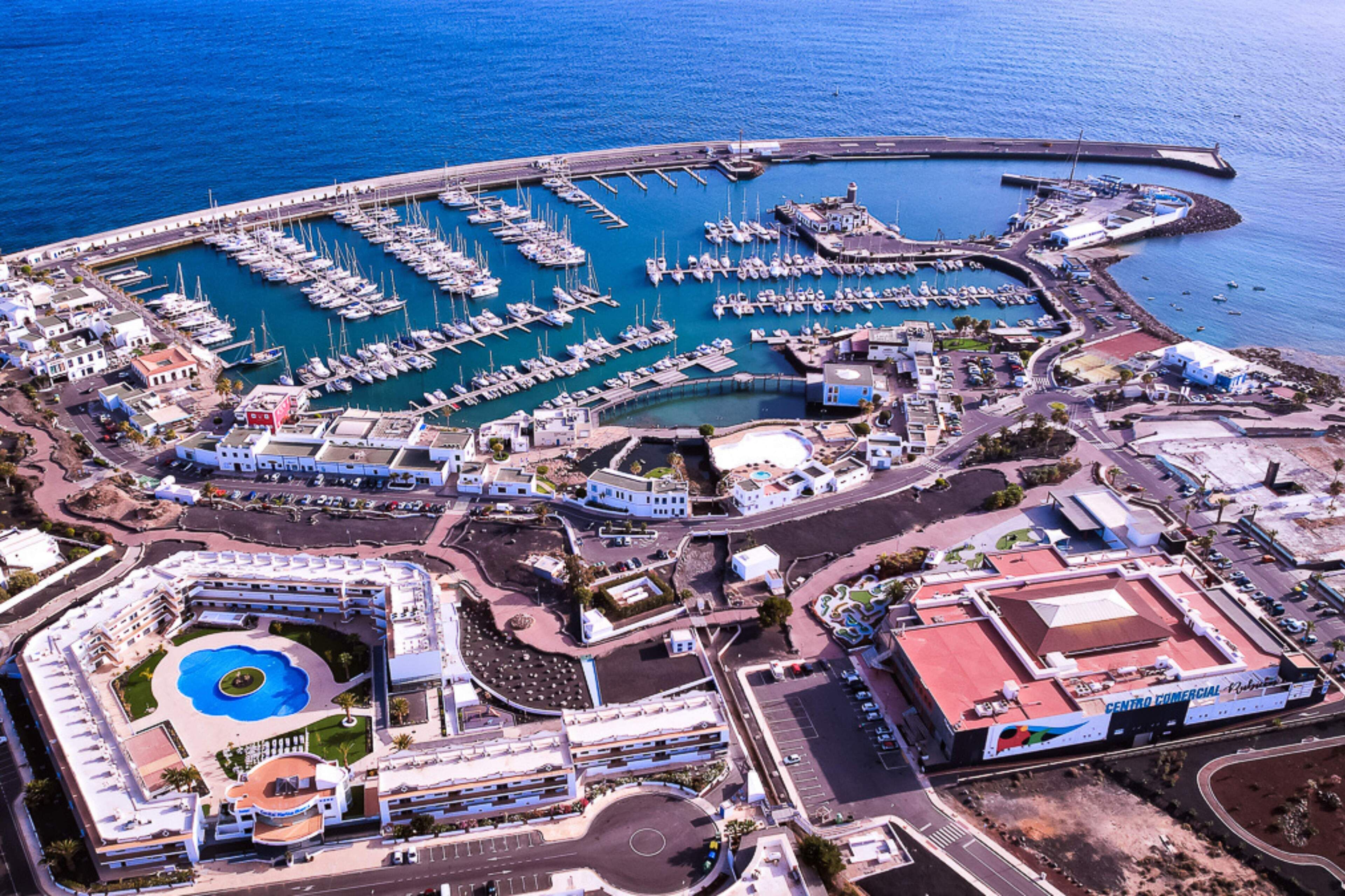 Vista aérea de un puerto deportivo con numerosas embarcaciones amarradas, una piscina central y edificios circundantes, incluyendo un centro comercial.