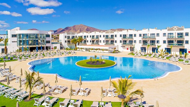 Um resort com uma grande piscina central, várias varandas e montanhas ao fundo.