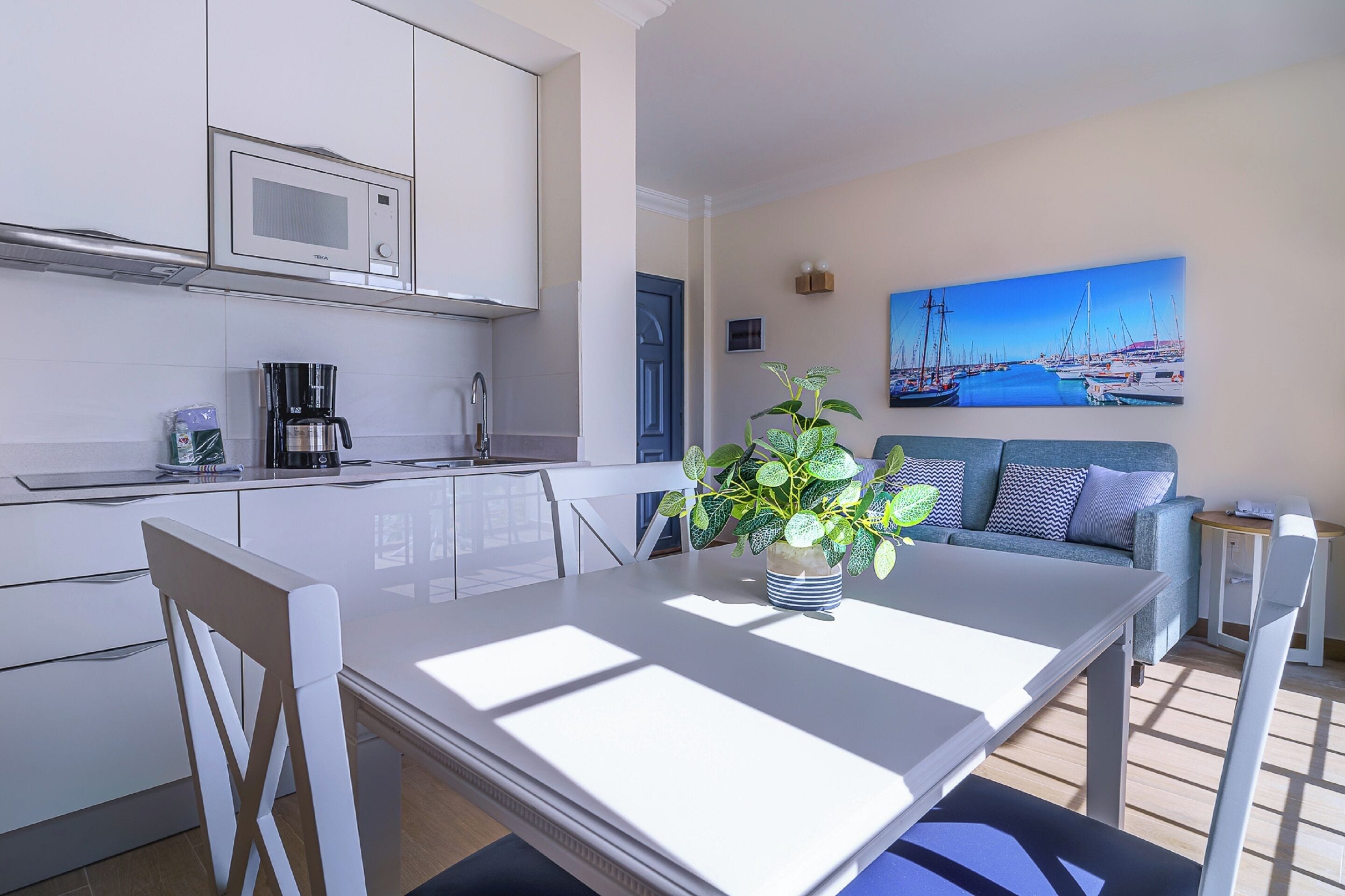 A modern kitchen with white cabinets, a microwave, and a coffee maker. There is a dining table with chairs and a potted plant. A sofa and a painting of a harbor are visible in the background.