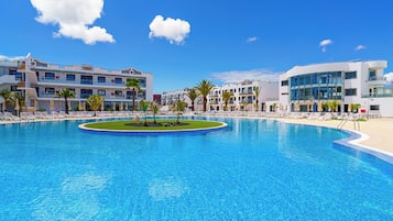 Una gran piscina con una isla central, rodeada de sillas de descanso y palmeras, con edificios modernos al fondo.