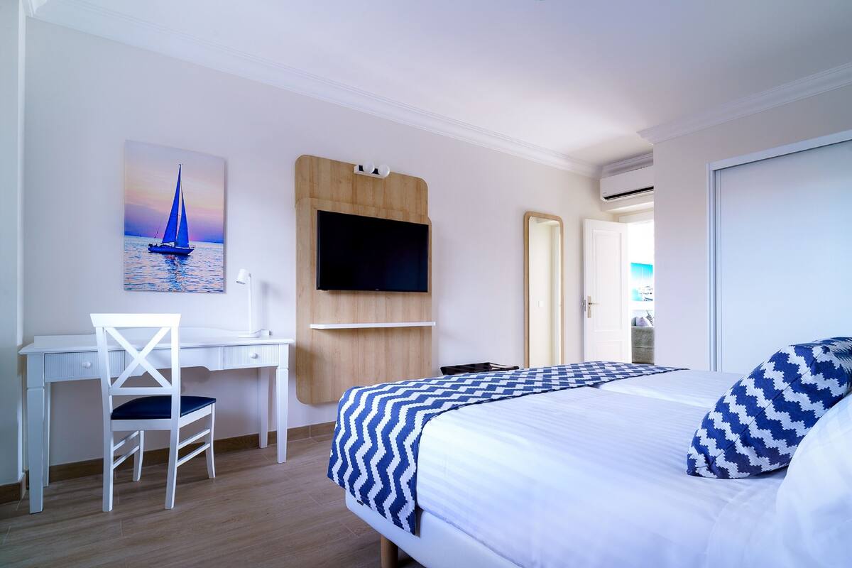 A hotel room with a bed, a desk, a chair, a TV mounted on a wooden panel, and a door leading to another room.