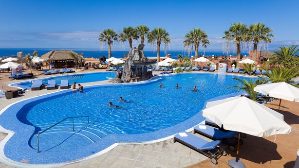 A swimming pool with a waterfall feature, surrounded by lounge chairs and umbrellas, with a view of the ocean. - Grand Hotel Callao (Adeje)