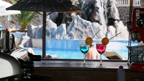 A bar area with two cocktails and a Coca-Cola machine, overlooking a pool with rocks and a thatched umbrella. - Grand Hotel Callao (Adeje)