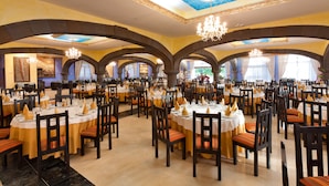 A large dining hall with multiple round tables, each set with white tablecloths and gold napkins. The room features arched ceilings and chandeliers. - Grand Hotel Callao (Adeje)