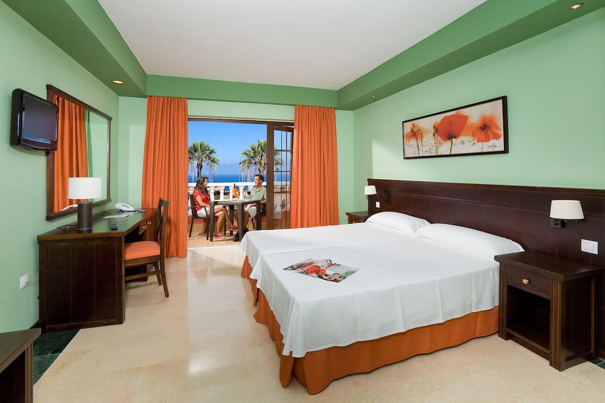 Habitación de hotel con cama, escritorio, silla y televisor. Posee un balcón con vistas a palmeras y al océano.