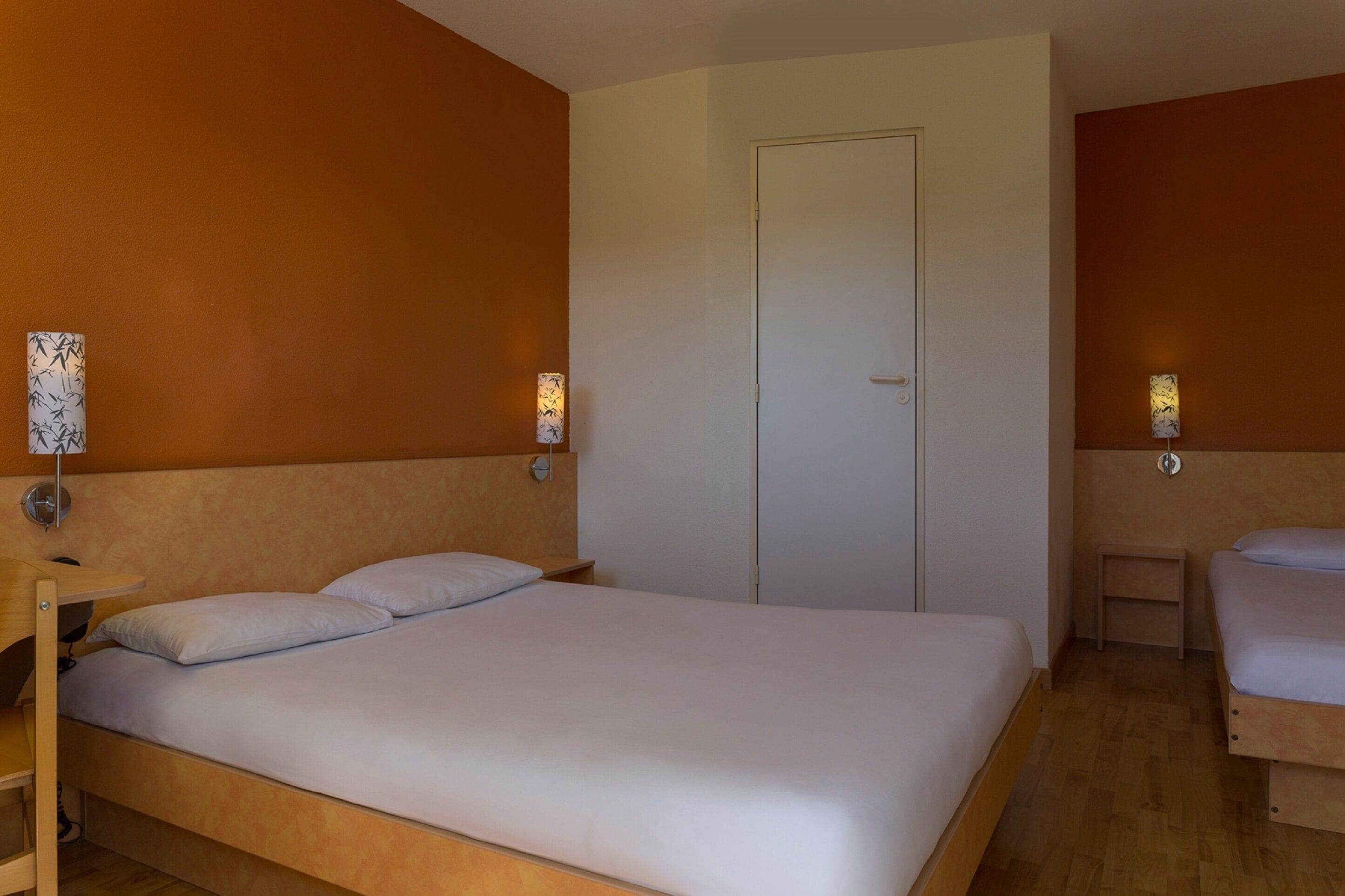 A single-bed room with a wooden headboard, a bedside table, and a nightstand. There is a door in the center of the room.