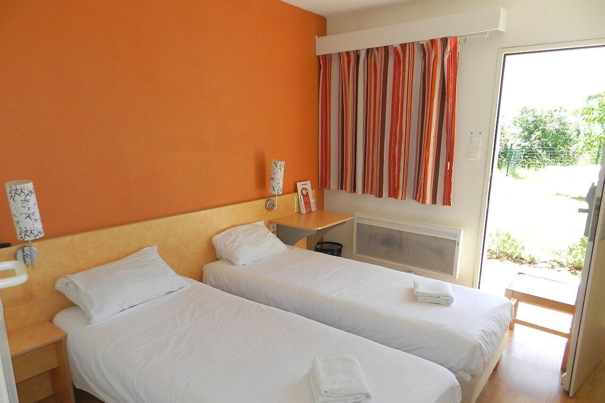 A hotel room with two single beds, a wooden headboard, a small bedside table with a lamp, a window with striped curtains, and a view of greenery outside.