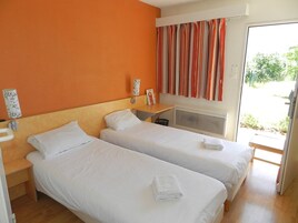 Quarto de hotel com duas camas de solteiro, cabeceira de madeira, mesinha de cabeceira com luminária, janela com cortinas listradas e vista para o verde lá fora.