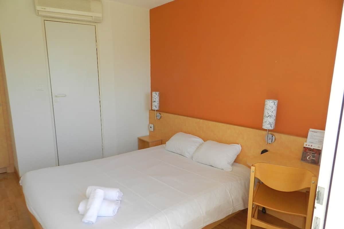 A neatly made bed with white linens and a folded towel, a wooden nightstand with a lamp, and an orange accent wall.