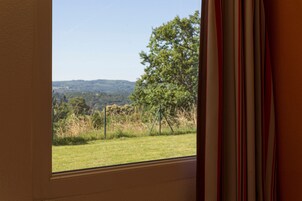 Janela com vista para um campo gramado, árvores e colinas ao longe.