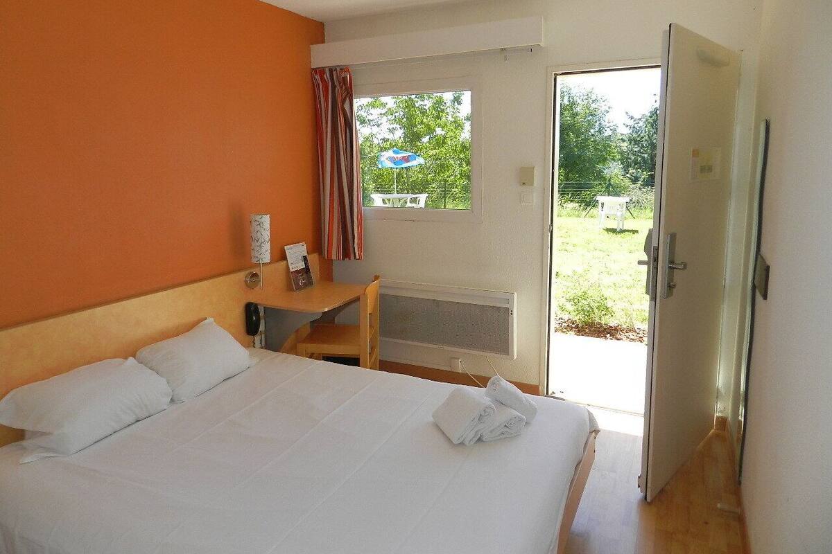 A hotel room with a bed, a desk, a chair, a window with curtains, and a door leading to an outdoor area.
