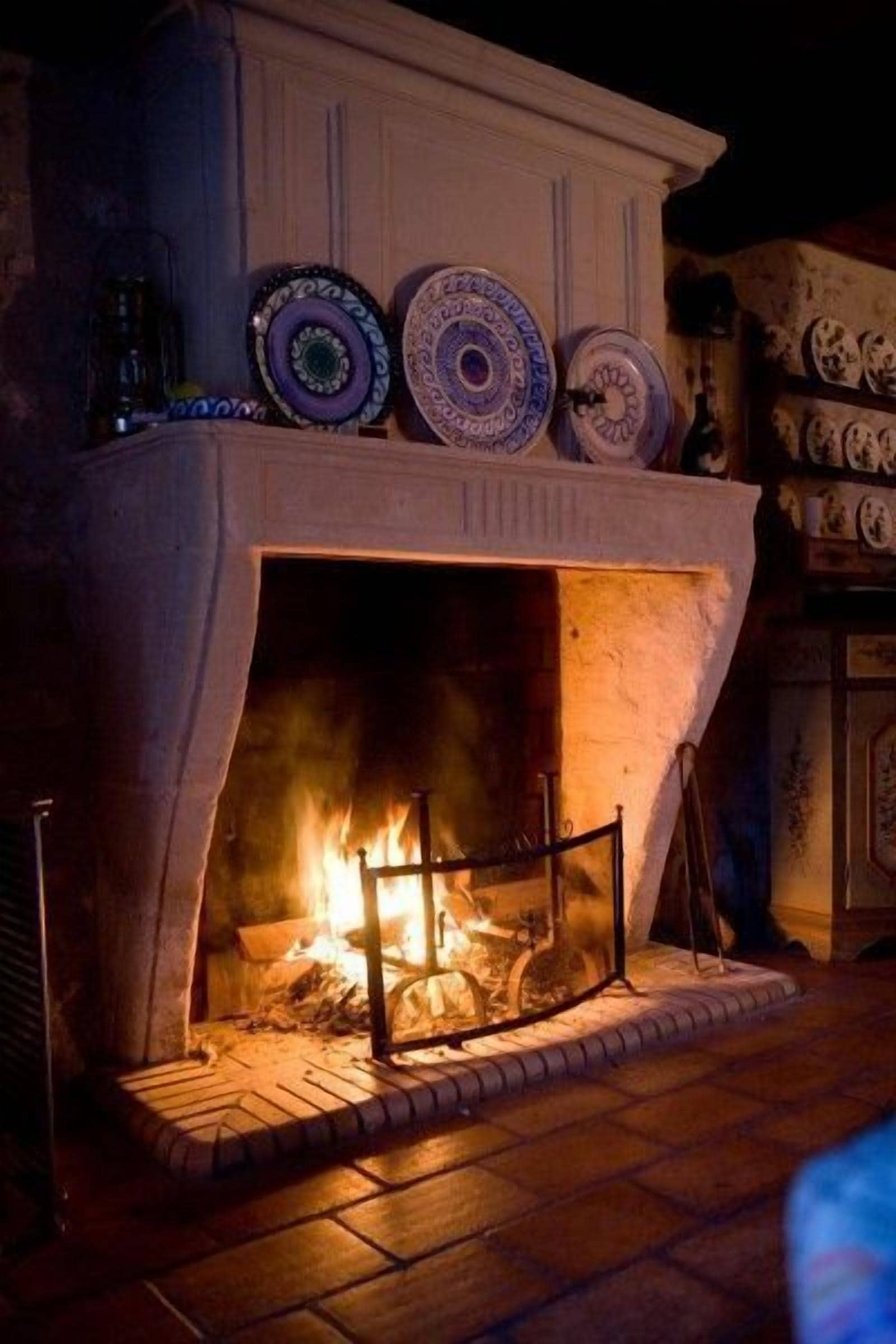 Una chimenea con platos decorativos encima, fuego encendido en su interior y suelo de ladrillo.