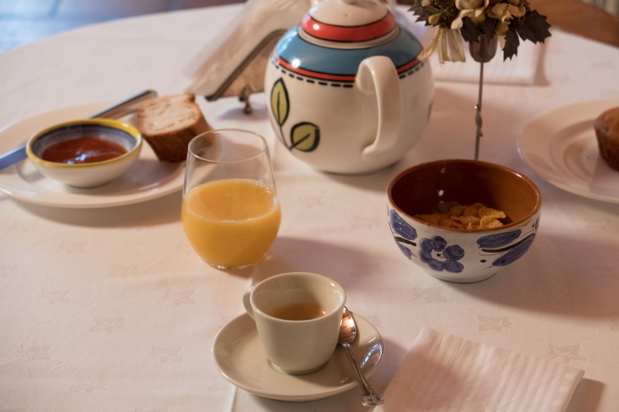 Ein Tisch mit einem Teekännchen, einer Tasse Kaffee, einer Schüssel mit Müsli, einem Glas Orangensaft und einem Teller mit Brot und Butter.