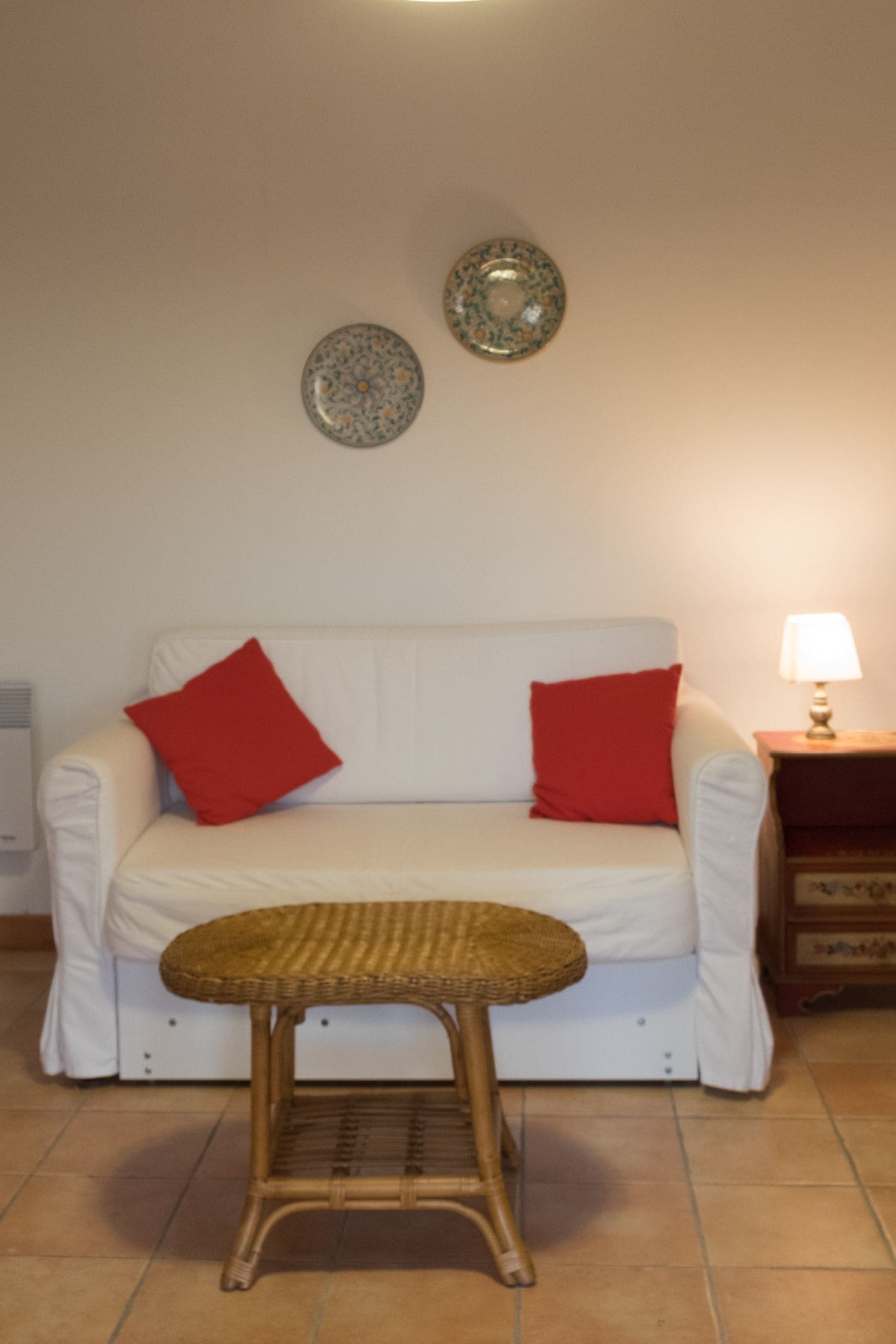 Un canapé blanc avec des coussins rouges, une chaise en osier, une petite table avec une lampe et deux assiettes décoratives accrochées au mur.