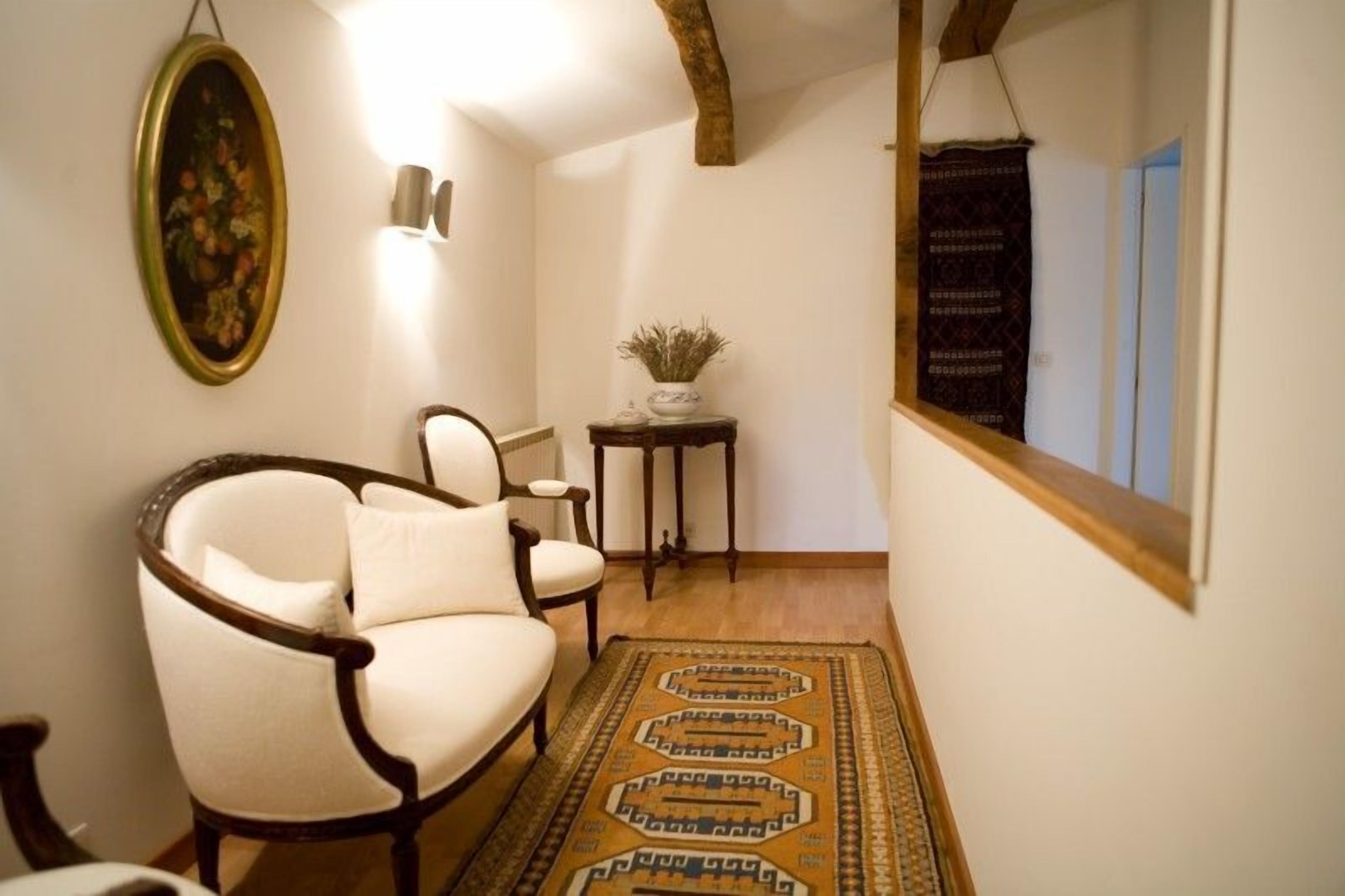 A narrow hallway with a wooden floor, a white armchair, a small table with a plant, and a framed painting on the wall.