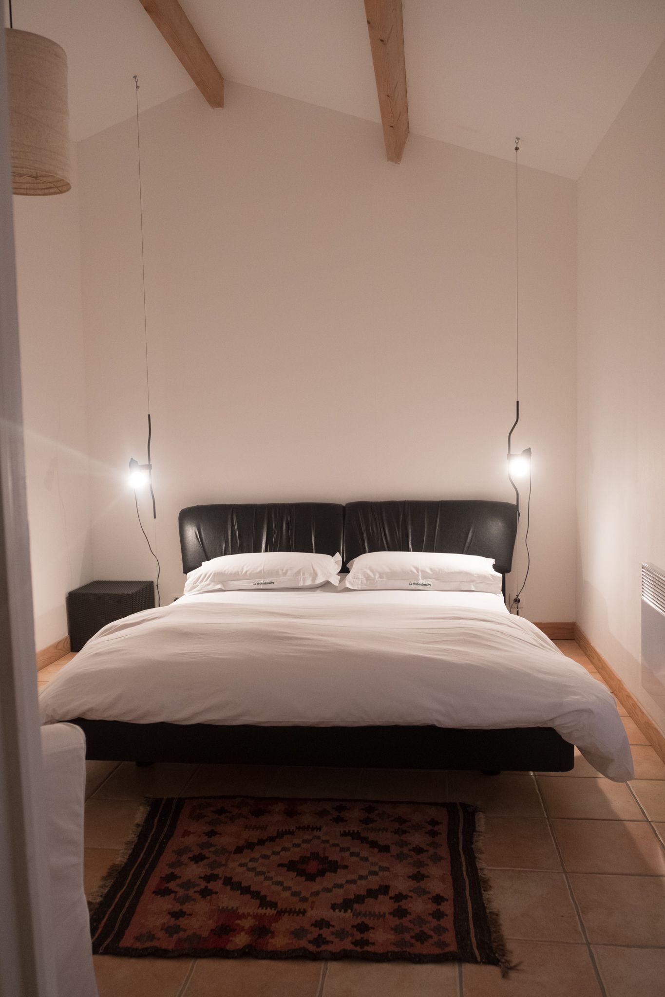 Quarto com cama, mesa de cabeceira, luminária na parede e tapete estampado.
