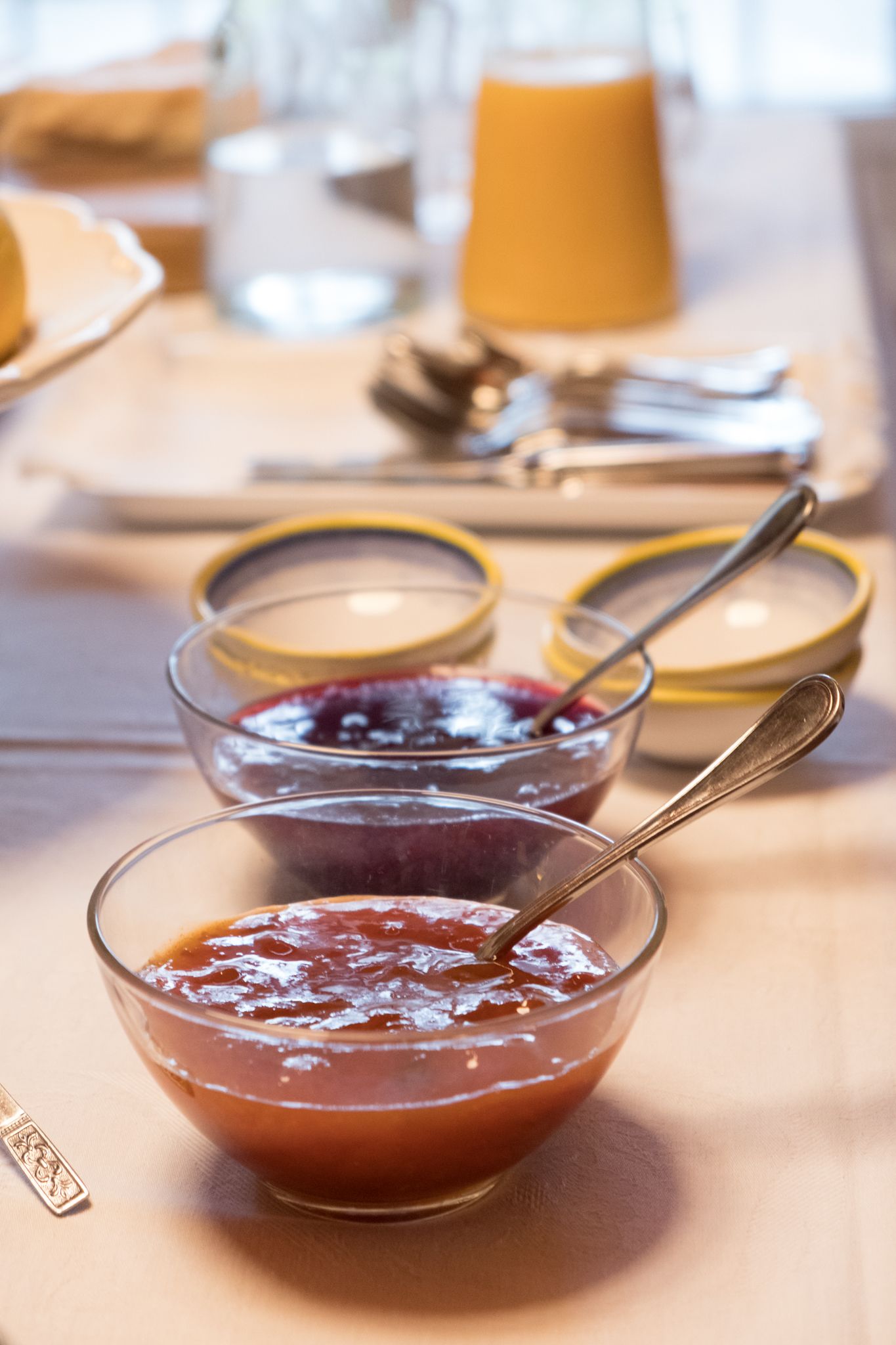 A table with three bowls of fruit preserves, a glass of orange juice, and a plate with a sandwich.