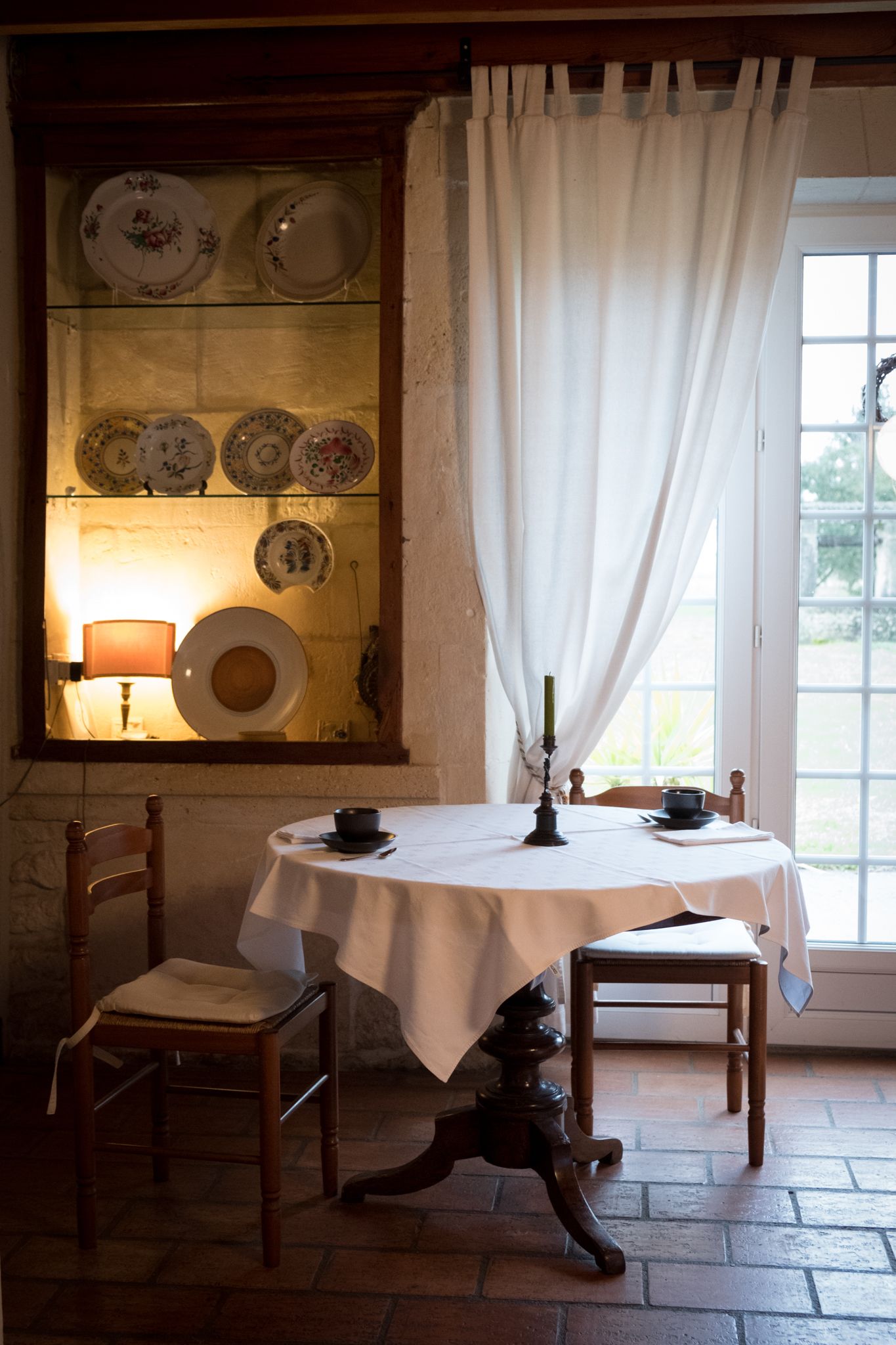 A dining area with a table set for two, a chair, a lamp, and a window with curtains.