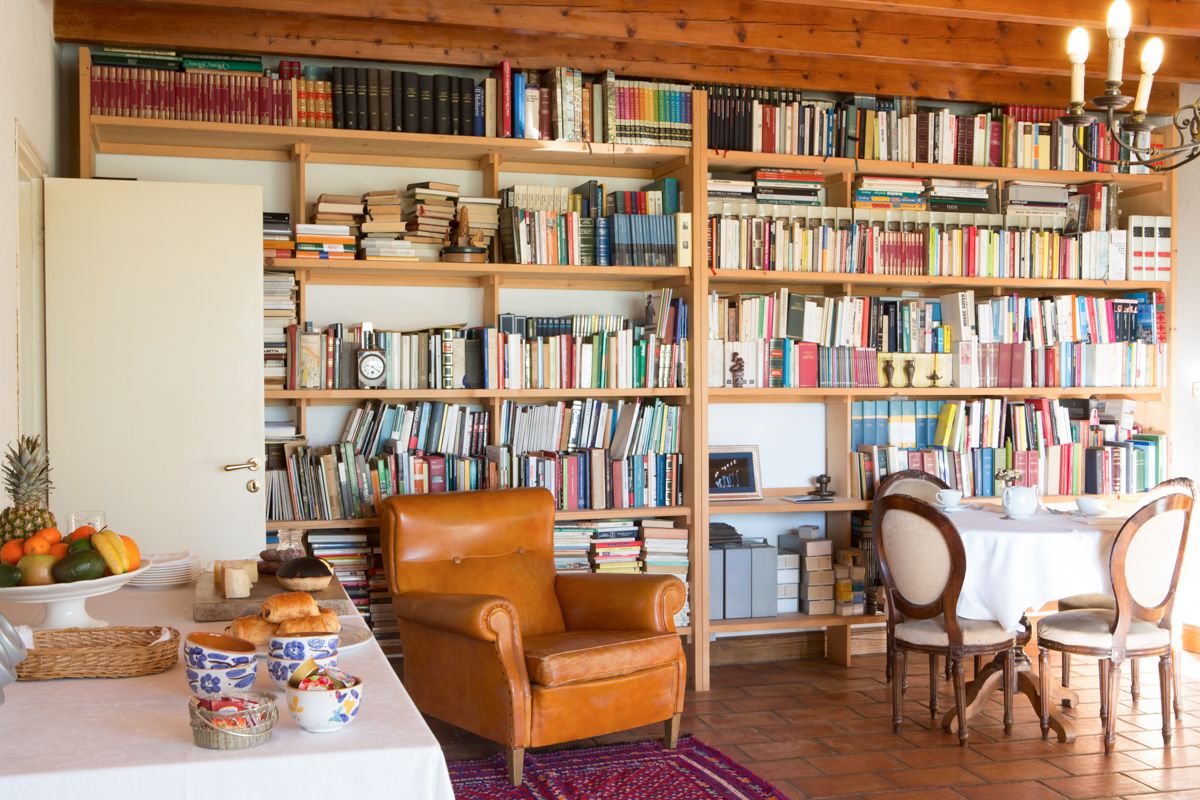 Una habitación acogedora con una estantería de madera llena de libros, un sillón cómodo, una mesa de comedor con sillas y un frutero a un lado.