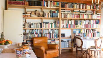 Una habitación acogedora con una estantería de madera llena de libros, un sillón cómodo, una mesa de comedor con sillas y un frutero a un lado.