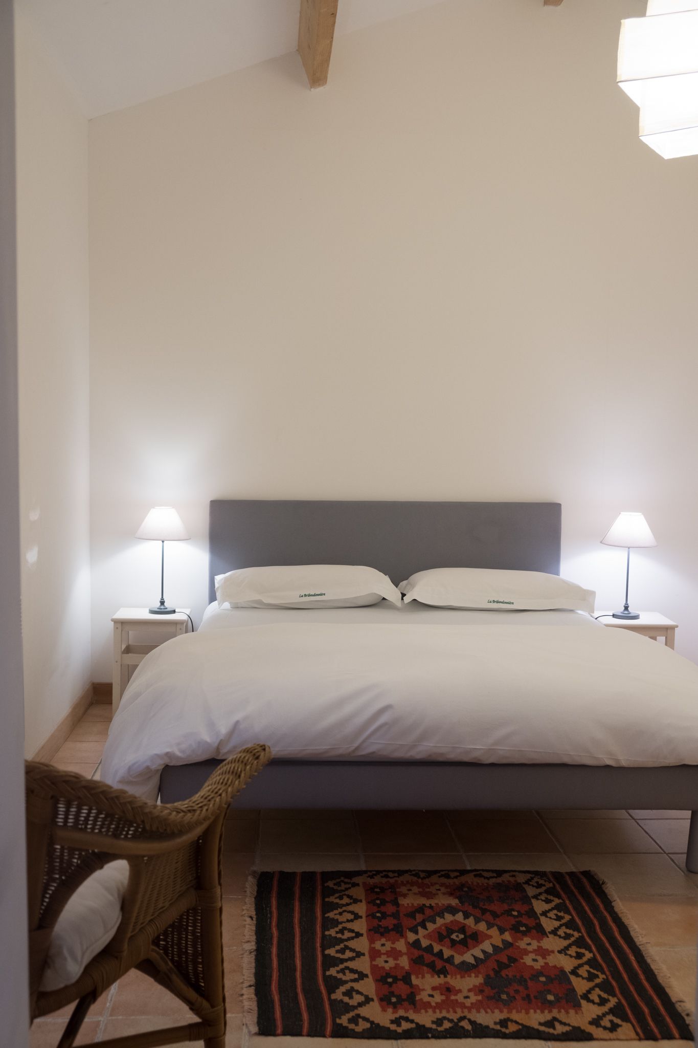 A bedroom with a bed, two bedside tables with lamps, a wicker chair, and a patterned rug.