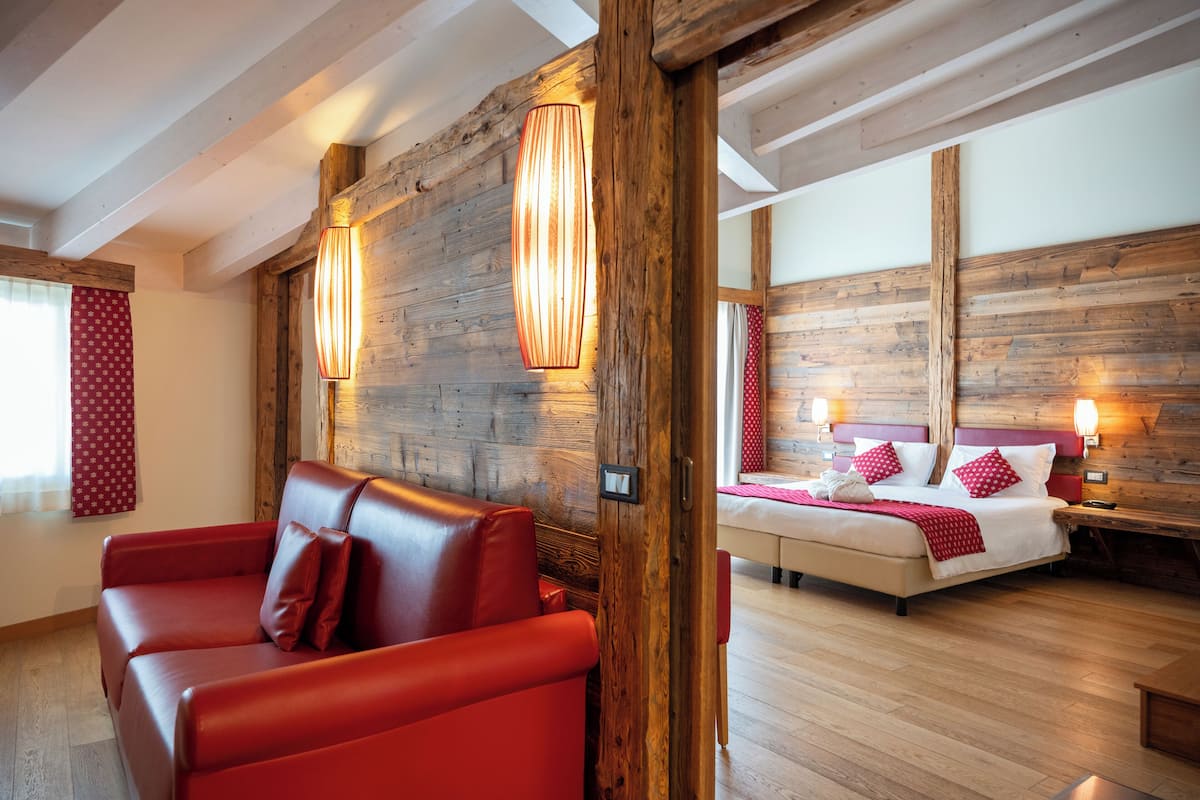 A room with a wooden wall, a bed with red pillows, a red sofa, and two wall-mounted lamps.