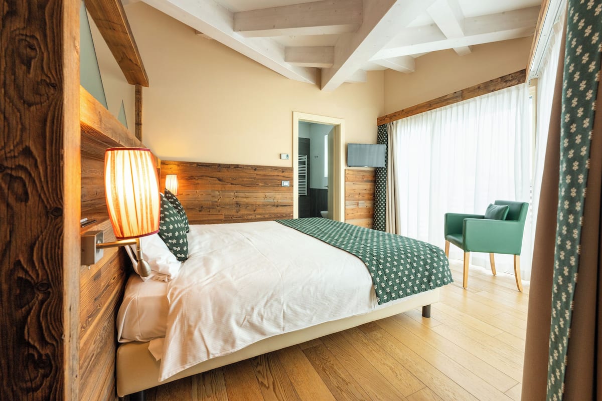 A bedroom with a wooden headboard, a bed with white and green bedding, a bedside lamp, a green chair, and a view of a balcony with curtains.