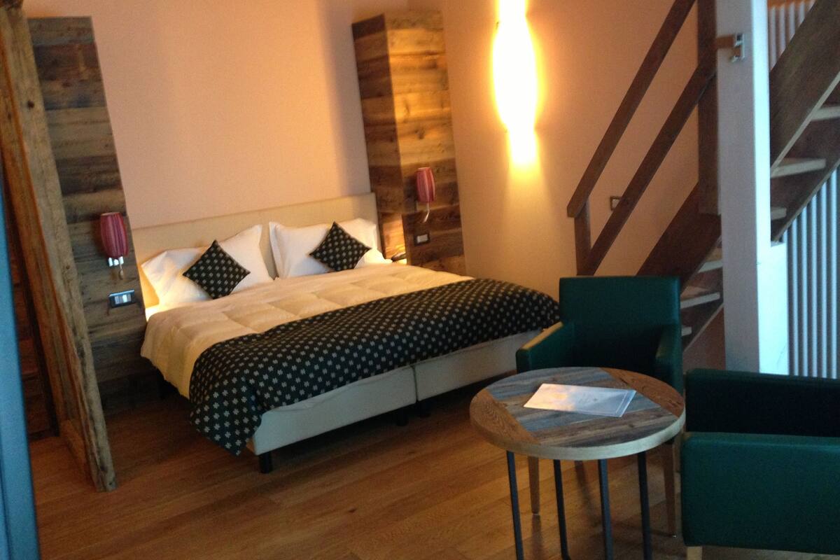 A bedroom with a bed, a chair, a small table, a staircase, and a wall-mounted light.