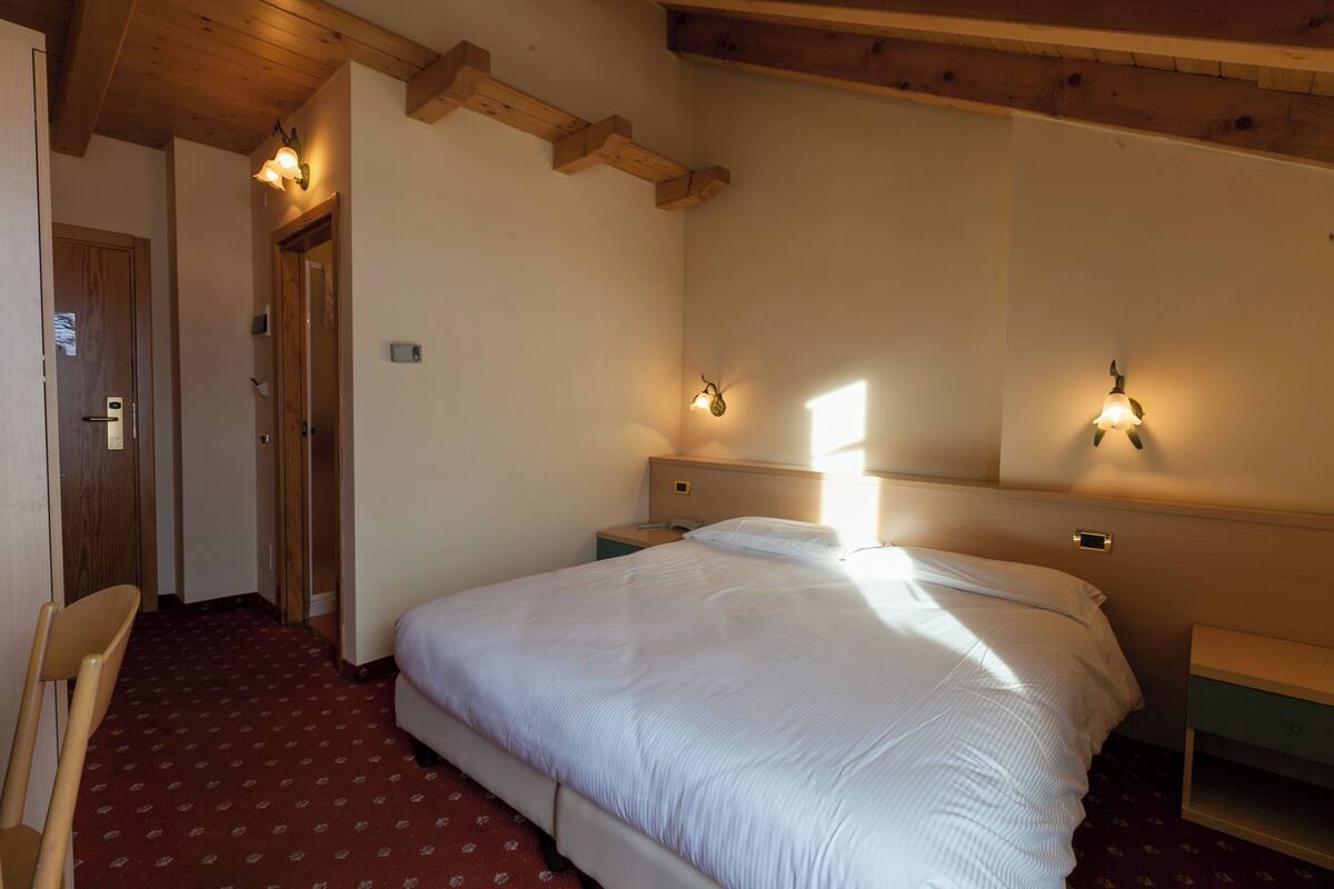 A hotel room with a bed, wooden ceiling beams, two bedside lamps, a nightstand, and a chair.
